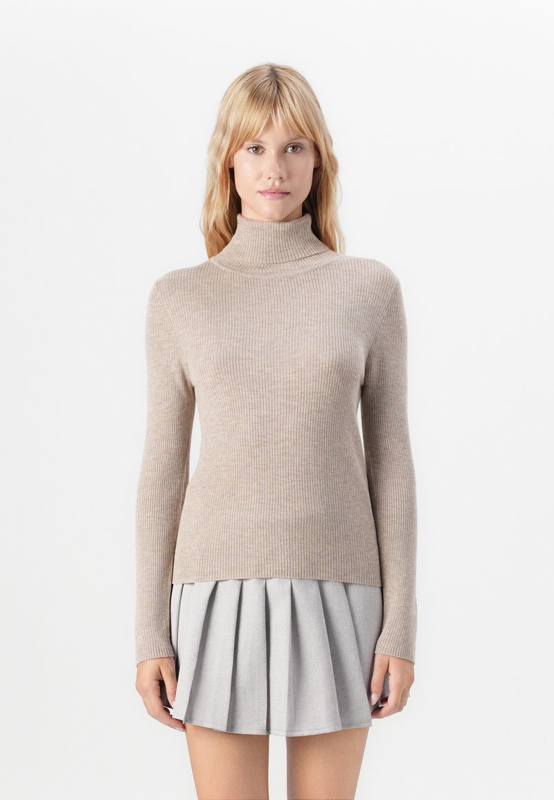 Kvinde iført en beige ribbestrikket turtleneck-sweater og en lysegrå plisseret mini-nederdel, stående foran en ensfarvet hvid baggrund.