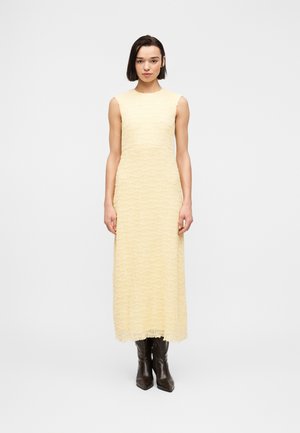 Vrouw die recht vooruit staat, gekleed in een mouwloze, lichtgele maxi-jurk met textuur en donkerbruine puntige laarzen, op een witte achtergrond.