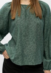 Langarmige groene blouse met textuur en pofmouwen, gedragen met zwarte broeken, hand in de zak, en steil bruin haar zichtbaar.