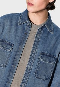Camicia in denim di blu sbiadito, con botones anteriori, colletto a punta e due tasche sul petto. Indossata sopra un maglione grigio chiaro a trama.
