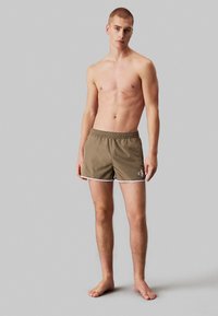 Pantaloni da bagno marroni con bordi bianchi, vita elastica e logo sull'orlo. Il modello sta in piedi scalzo su uno sfondo neutro, mostrando la vestibilità.