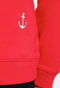 Červená mikina s texturovaným povrchem, na rukávu s malou bílou vyšívanou kotvou a žebrovanými manžetami na spodním okraji.