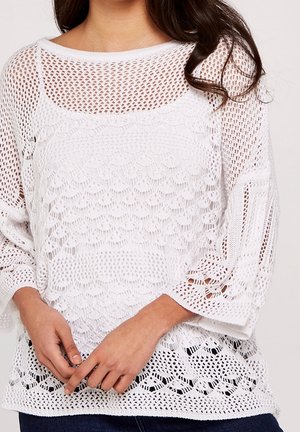 Kvinde iført en hvid, langærmet hækletrøje med indviklede åbne mønstre, kombineret med mørkeblå jeans.