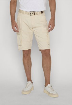 Man draagt beige cargoshorts met meerdere zakken, een beige riem, witte sneakers en een wit T-shirt, staand tegen een effen achtergrond.
