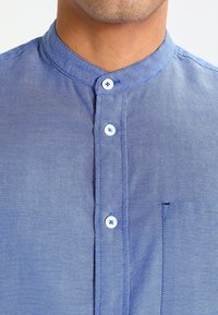 Jasnoniebieska, teksturowana koszula z kołnierzykiem stójkowym, wyposażona w białe guziki, kieszeń na piersi i widoczne detale szycia.