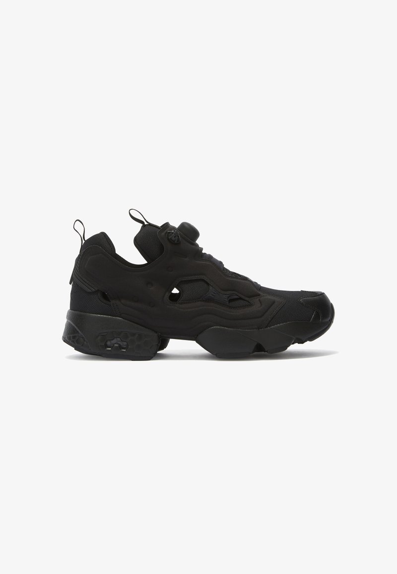 Sapatilhas pretas desportivas com um upper texturizado, detalhes hexagonais na sola, e um design slip-on com um fecho central para ajustamento.