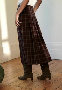 Jupe maxi en tartan bordeaux avec des plis, associée à des bottes en suédine vert olive hauteur genoux avec une fermeture éclair sur le côté. Fond en parquet en bois.