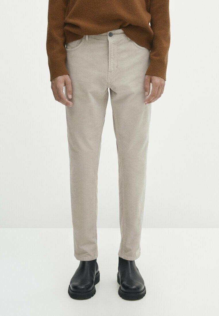 Personne portant un pantalon beige droit, un pull marron et des bottines noires, debout devant un fond clair uni.