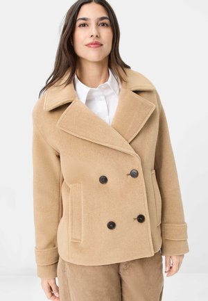 Frau trägt einen beigefarbenen, doppelt geknöpften Wollmantel über einem weißen Hemd und braunen Hosen, steht vor einem schlichten weißen Hintergrund.
