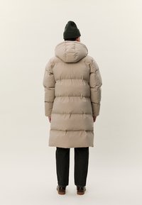 Άτομο που φοράει ταμπά επενδυμένο παλτό με κουκούλα, μαύρο σκουφάκι, σκούρο παντελόνι και καφέ παπούτσια, στέκεται με την πλάτη προς το μέρος μας, μπροστά σε ένα απλό φόντο.