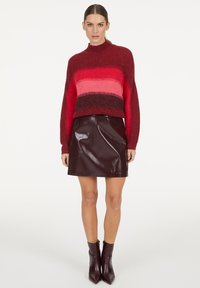 Frau in einem rot-rosa gestreiften Strickpullover, glänzendem dunkelburgunderfarbenen Miniröckchen und passenden spitzen Knöchelstiefeln, die vor einem weißen Hintergrund steht.