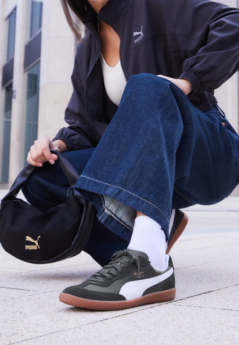 Personne accroupie en extérieur portant des baskets Puma noires et blanches, des chaussettes blanches, un jean bleu, une veste Puma noire, tenant un sac Puma noir.