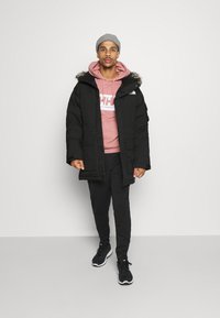 Zwarte gewatteerde jas met een capuchon van bont, gedragen over een roze hoodie en zwarte broek. Model draagt een grijze beanie en zwarte sneakers.