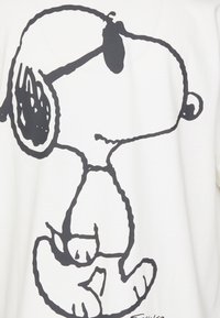 T-shirt blanc présentant un dessin au trait noir d'un chien cartoon avec des oreilles tombantes et un corps arrondi, se tenant sur deux pattes.