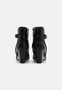 Para czarnych skórzanych botków na kostkę na masywnym obcasie, z krzyżującymi się paskami i złotymi klamrami, widzianych od tyłu na białym tle.