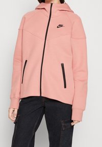 Rosa Kapuzenjacke mit Reißverschluss aus weichem Stoff, mit Kapuze, schwarzen Akzenten, seitlichen Reißverschlusstaschen und einem kleinen schwarzen Logo auf der Brust.