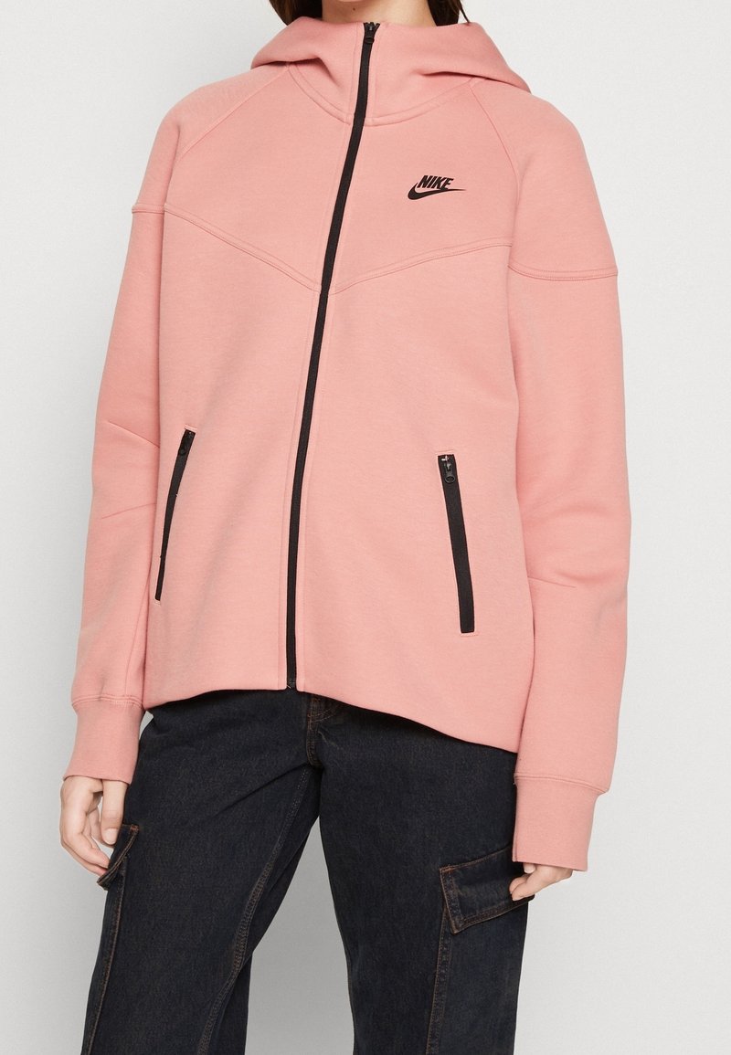 Rosa Kapuzenjacke mit Reißverschluss aus weichem Stoff, mit Kapuze, schwarzen Akzenten, seitlichen Reißverschlusstaschen und einem kleinen schwarzen Logo auf der Brust.