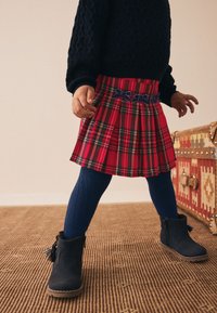 Röd rutig plisserad kjol med en blå rosett, kombinerad med marinblå stickade strumpbyxor och svarta ankelboots. En texturerad tröja kompletterar outfiten.