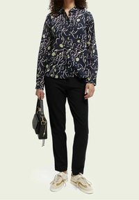 Marineblå skjorte med knapper og et abstrakt mønster i pink og grøn, parret med sorte bukser og beige sneakers. Sort håndtaske med guld accenter.