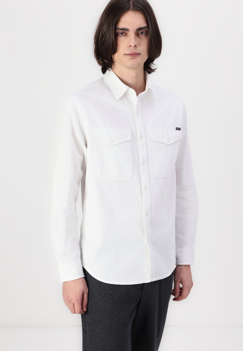 Langarmige witte button-up shirt van lichte stof, met twee borstzakken en een afgeronde zoom, gecombineerd met donkergrijze broek.