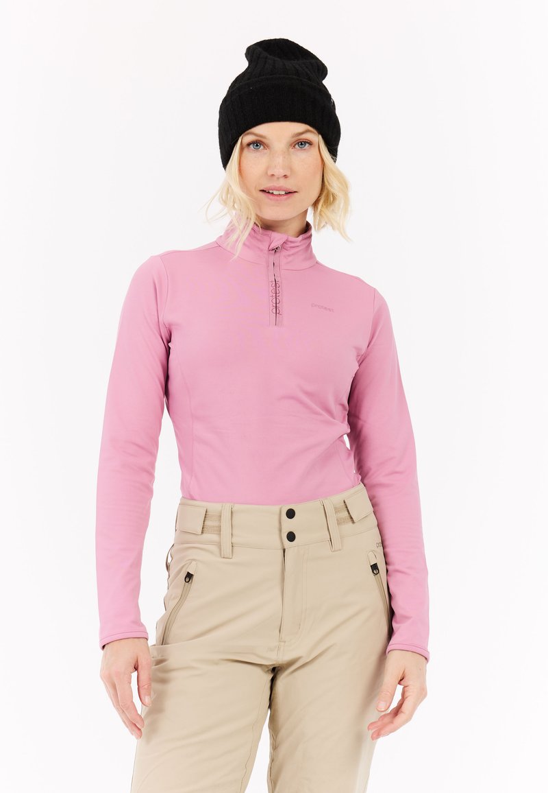 Ilga rankovė, šviesiai rožinė palaidinė su pusiau užtrauktuku ir logotipu, derinama su smėlio spalvos kelnėmis su kišenėmis ir juodais sagomis. Juodas megztas skrybėlė.