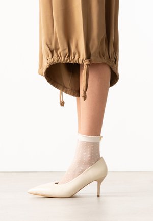 Benen met doorschijnende witte enkelsokken met stippen en crèmekleurige puntige hoge hakken, met een bruine rok met een verstelbare tunnelkoordzoom.