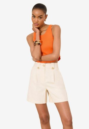 Frau trägt ein ärmelloses oranges Oberteil und hoch taillierte cremefarbene Shorts mit goldenen Knöpfen, posiert mit einer Hand am Kinn.