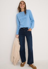 Sweatshirt bleu clair avec texte en relief, jeans bleu foncé, veste en cordon beige ample et baskets marron. Textures lisses et design décontracté.
