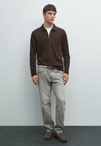Ung mand iført en brun langærmet polo, lysegrå jeans med et brunt bælte og brune ruskinds loafers, der står ved en lys væg.