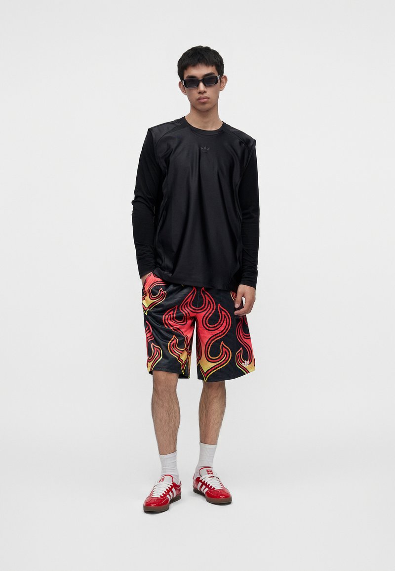Giovane uomo che indossa una maglia nera a maniche lunghe, pantaloncini rossi e gialli con motivo a fiamme, calzini bianchi, scarpe da ginnastica rosse e occhiali da sole neri su uno sfondo semplice.