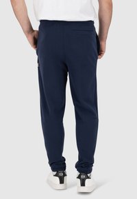 Pantaloni jogger blu navy con una superficie liscia, cintura elastica e polsini affusolati; presentano una tasca posteriore e un branding discreto sul lato.