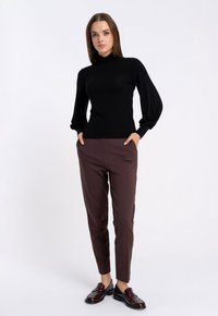 Czarny długi rękaw z wysokim kołnierzem i bufiastymi rękawami, w połączeniu z dopasowanymi brązowymi spodniami i bordowymi mokasynami. Białe tło.