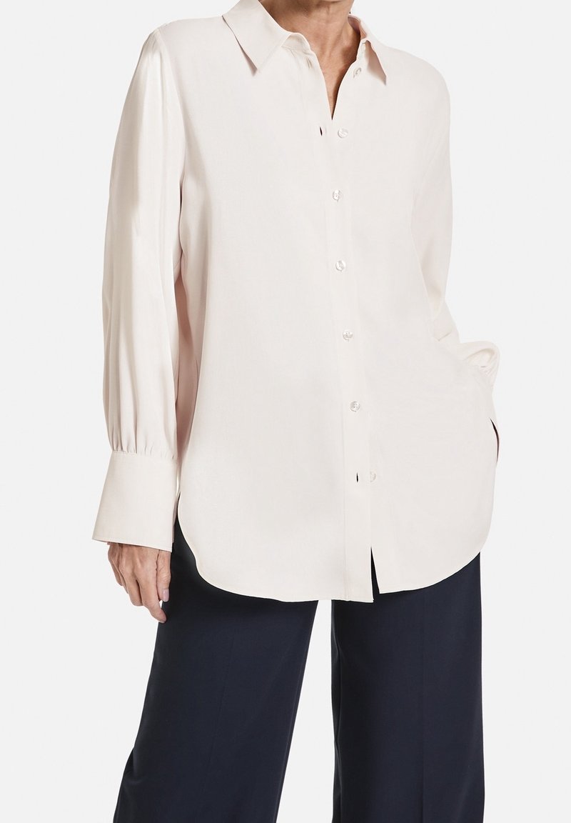 Chemise boutonnée rose clair avec un col, des manches longues et un ourlet arrondi. Tissu lisse avec un léger éclat et des manches à revers.