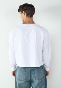 Biała bluzka z długim rękawem o skróconym kroju i okrągłym dekolcie, zestawiona z niebieskimi, wysokimi dżinsami z przetarciami.