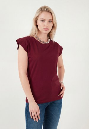 Naine blondide juustega, seljas burgundia lühikeste varrukatega pluus, sinised teksad ja hõbedane helmeke kaelakee, seisab ühevärvilise tausta ees.