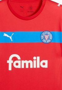 Rød trøje med en blå vandret stribe og et hvidt logo med teksten "familia." Den har rund hals og Puma-logoet på venstre skulder.