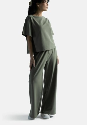 Nainen yllään löysä oliivinvihreä paita ja leveälahkeiset housut valkoisilla lenkkareilla, katsoen sivulle yksiväristä taustaa vasten.