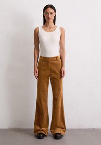 Witte mouwloze bodysuit gecombineerd met wijdvallende, gestructureerde bruine corduroy broek en zwarte platform schoenen, staand op een grijze vloer.