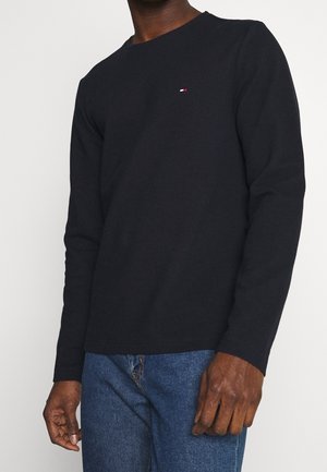 Schwarzes Langarmshirt aus strukturiertem Stoff mit rundem Ausschnitt und kleinem roten, weißen und blauen Logo auf der Brust. Kombiniert mit blauen Jeans.