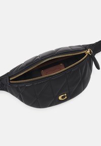 Banane en cuir noir matelassé avec fermeture éclair dorée partiellement ouverte, montrant une étiquette en cuir marron avec la marque à l'intérieur et un logo doré « C » sur le devant.