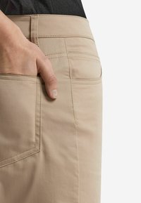 Beige Hose mit glatter Textur, ausgestattet mit einer Gesäßtasche und sichtbaren Nähten. Eine Hand ruht in einer Seitentasche.