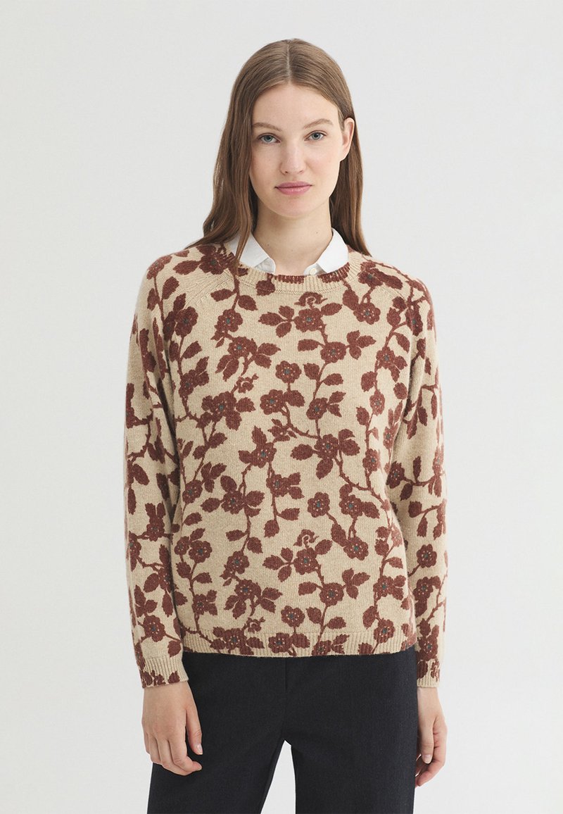 Jeune femme portant un pull beige avec un motif floral marron et une chemise blanche à col en dessous, debout devant un fond clair uni.