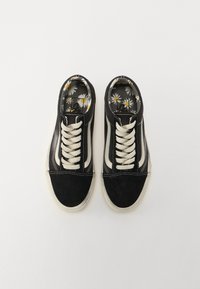 Une paire de baskets noires et blanches avec des lacets blancs, une semelle blanche et des semelles intérieures à motif floral de marguerites sur un fond blanc uni.