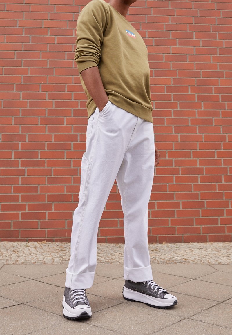 Sweat-shirt vert olive avec logo, pantalon ample blanc avec poches latérales, et baskets à plateforme noir-gris avec accents blancs. Fond en brique.