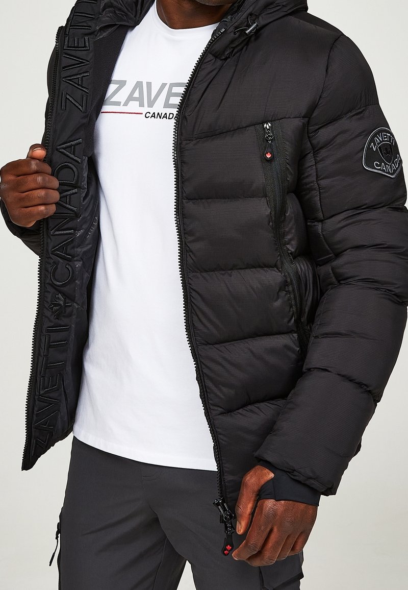 Schwarze Steppjacke mit Reißverschlüssen, strukturierter Stoff, verstellbarer Kapuze und Logopatch. Getragen über einem weißen T-Shirt mit gedrucktem Markennamen.