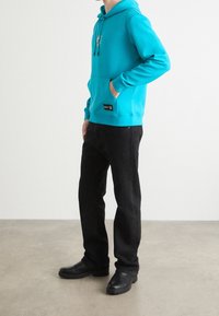 Person som bär en turkos hoodie med framficka och svarta jeans, står på en grå golv framför en enkel vit vägg.