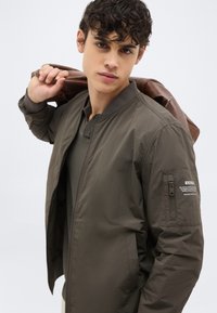 Νέος άνδρας με σκούρα κατσαρά μαλλιά, φορώντας ένα πράσινο χακί μπουφάν τύπου bomber και κρατώντας μία καφέ δερμάτινη τσάντα στον ώμο του.