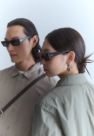 Juodos stačiakampės saulės akiniai su blizgiu paviršiumi, ant čiaupo esanti Ray-Ban logo, vilkintys du žmonės su subtiliais drabužiais.