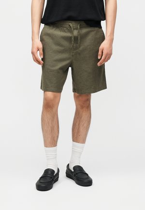 Mężczyzna w oliwkowozielonych szortach ze sznurkiem, białych skarpetkach za kostkę, czarnych butach wsuwanych i czarnym t-shircie, stojący na jasnym tle.