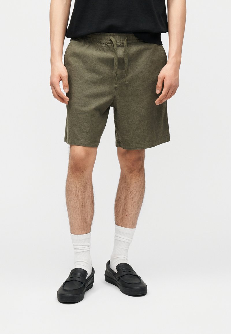 Άνδρας που φοράει ελαφρώς χακί σορτς με κορδόνια, λευκές κάλτσες μέχρι τον αστράγαλο, μαύρα παπούτσια τύπου slip-on και μαύρο μπλουζάκι, στέκεται σε ανοιχτό φόντο.